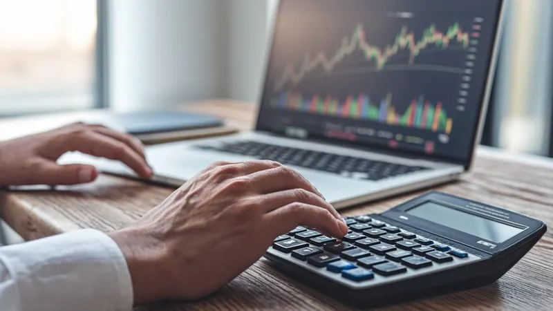 Hands using a calculator beside a laptop showing a financial chart